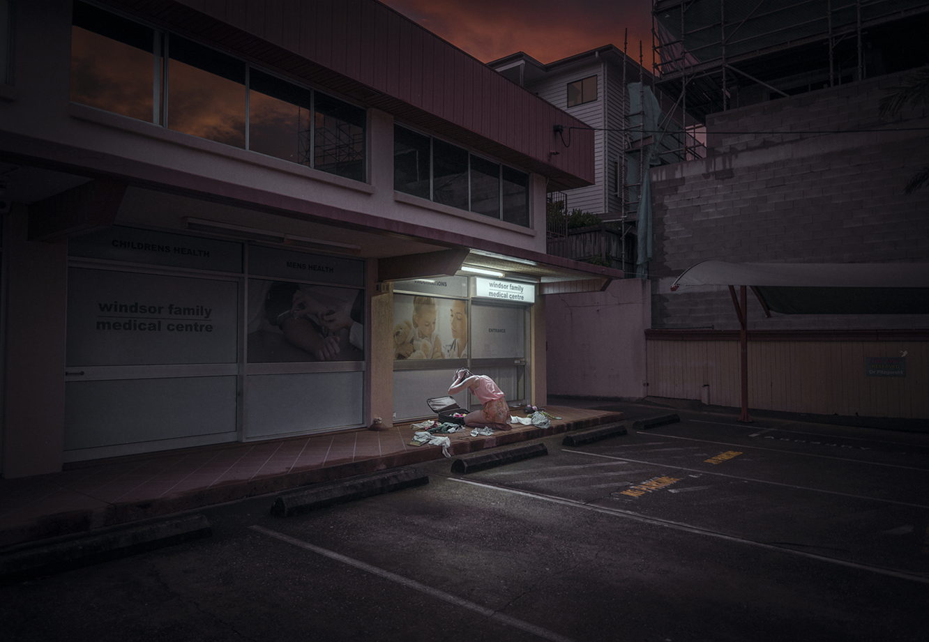 picture of a woman sitting on the side walk with her hands on her head, looking at an opened messy suitcase, outside of a storefront that says "windsor family medical centre"