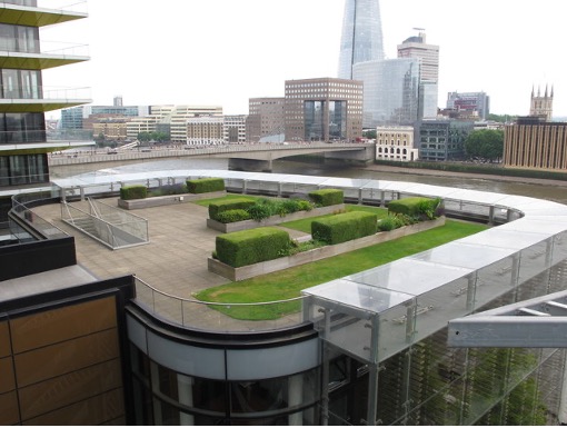 A modern rooftop garden with neatly trimmed hedges and green spaces overlooks London's skyline.