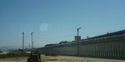 The picture shows a small tractor driving through a sandy field on a sunny day. On the right side of the picture you can see a long wall with barbed wire on top of it. In the background there are buildings of a city.