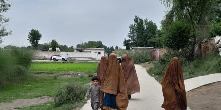Four women wearing burkas walk along a path next to a patch of grass, with residential buildings in the background. One woman holds a child's hand.