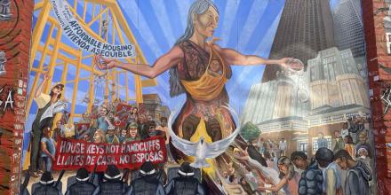 Photo of a street art wall picturing a woman standing tall over a group of people holding a sign that says "house keys not handcuffs", with a dove flying in the forefront and a line of police officers in riot gear facing towards the woman. In the background, a structure holds a sign that says "affordable housing" over a blue sky and skyscrapers in the background