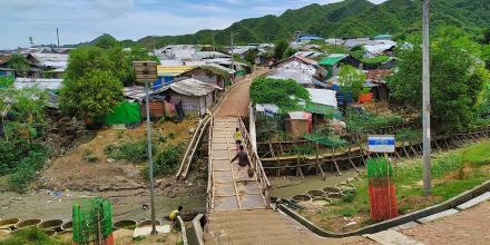 picture of a camp, with barracks in the back of the picture, and a wooden bridge in the middle, with people walking across it carrying bags of food