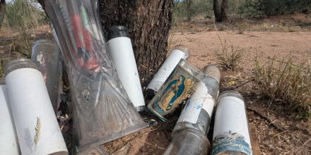 Empty glass containers, some holding the remains of candles and some bearing the image of the Virgin Mary, are piled at the base of a tree trunk