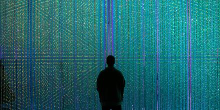 A person stands in front of a huge screen with vertical blue and purple lines of light 