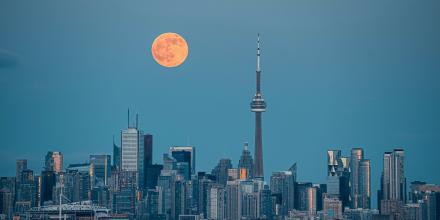 Toronto skyline as seen from the water. A full moon sits large and orange in the sky above the skyscrapers 