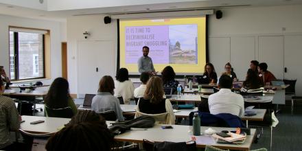 A man stands at the front of a seminar room smiling and gesturing. Others sitting at the panel and in rows listen. On the board behind him, a slide reads 'it is time to decriminalise migrant smuggling'