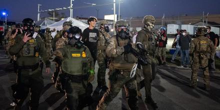 Federal border patrol agents wearing camo uniform and bullet proof vests walk along a street at night, some wearing heavy duty gas masks and some carrying large guns. A young man, smiling, is led by the arm by one of the agents. Protesters, wearing face masks or gas masks and goggles, stand at the edge of the street