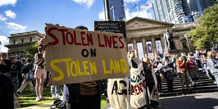 A person holds a sign reading 'Stolen lives on stolen land' with a group of protesters standing outside a building with colums on its facade – 