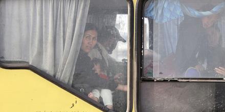 A woman sits next to a man – her and several children look out from the window of the yellow bus they are sat inside, with writing in Farsi on the outside of the bus