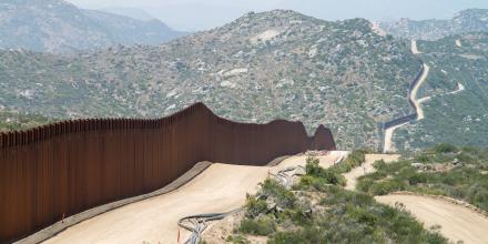 The US-Mexico border wall, brown against the green and sand scrubby hilly landscape, snakes over the hills