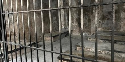Inside of what looks like an abandoned cell, as seen through the bars. Conditions look terrible, there are no people or furnishings inside. Everything is dirty