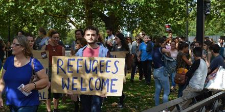 A man holds a cardboard sign reading Refugees Welcome. He stands under trees with other people around, some also holding cardboard placards