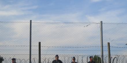 a few border guards stand in line behind multiple rounds of barbed wire fences, over a background with a blue sky. 