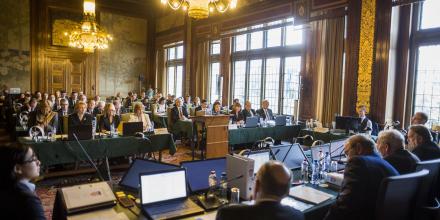 The inside of a court room with chandeliers and high ceilings. Several people sit with laptops and microphones at a table at the front of the room, facing rows of people in the main area of the room