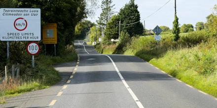 A road undulates over gentle bumps in the landscape. Green trees on the left and green fields on the right. On the left is a circular 100 km/hr sign, and above this, a much larger sign in English and Irish reading: speed limits kilometres per hour