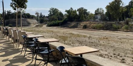 Tables and chairs are lined up along the edge of a brick paved street next to a wall. They overlook a dusty empty patch of land, with scrubby trees, barely distinguishable fence and watchtower in the background