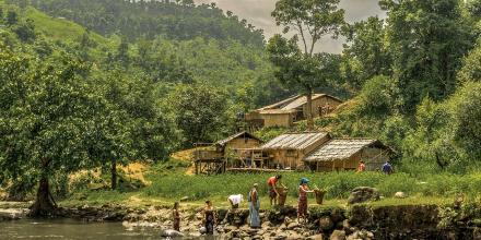 A lush green valley and river next to which people carry baskets. Small huts sit nearby at the foot of the hill