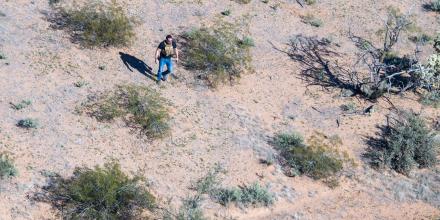A man wearing sunglasses, jeans, black t-shirt and a beige police vest walks alone in a desert area – brown earth and some green scrub 