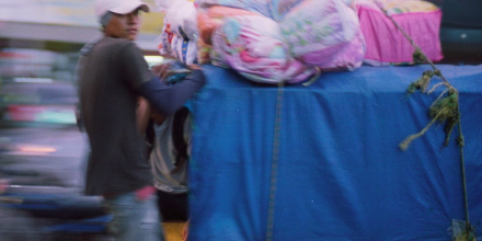 a person wearing a blue shirt and a white cap is holding on to the back of a truck, filled with things and secured with ropes that hold bags on top of it. The truck appears to be moving 
