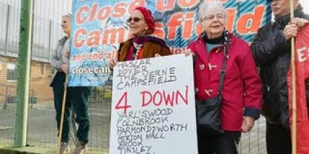 Several people stand in front of a fence holding a big banner reading 'Close down Campsfield'. Two people with a placard stand in front of the banner with a placard reading '4 down. 7 to go' and a list of the detention centres