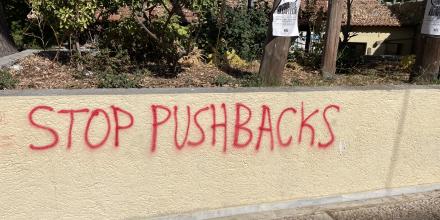 Red graffiti on a low, beige coloured wall reading 'stop pushbacks'. Above the wall are trees and a paper poster in Greek