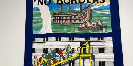A photograph of a tapestry hanging on a wall depicting people waving as they walk up a gangway onto a ship. It reads 'Justice knows no borders' at the top and 'Dorset' at the bottom