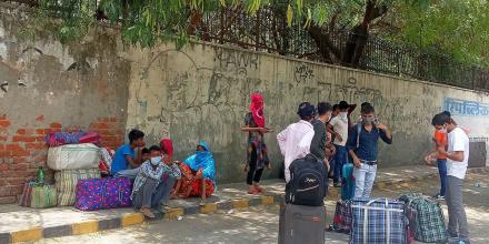 People wearing covid masks stand around with large suitcases next to a worn out bus. Some sit on the pavement next to a graffitied wall 