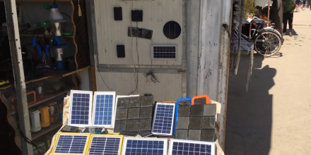 Small solar chargers laid out on a table under a shelter next to a dusty pathway in a refugee camp