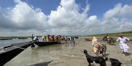 People get on and off a narrow boat moored at the edge of a river, against clouds, blue sky and green banks. A man holding a staff waits. Two goats are tethered to another mooring line. A bicycle waits for its owner