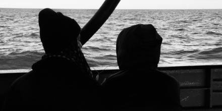 Two people look out to sea from on board a boat in a grayscale scene. They share a wire set of headphones, which hang between their figures against the backdrop of waves