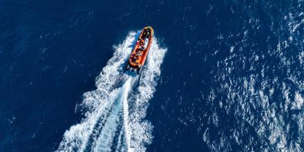 An orange lifeboat speeds through the ocean, as seen from above. At a distance, a white body bag can be seen on board