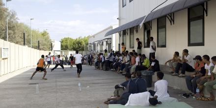 Men sit on mats and on concrete benches around the edge of a small concrete yard while others kick a ball around. Colours are grey and pale white, institutional. Except for the green of the mat and orange of the vest of a player