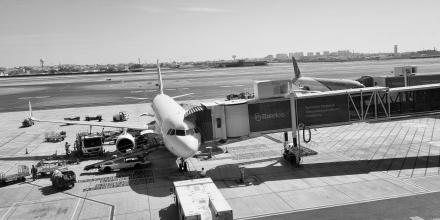 a black and white image of a plane parked at a big airport with a boarding tunnel attached, as seen through a window from the airport building 