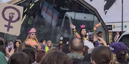 People stand in a crowd watching a woman wearing bright colours stood on a stage or podium, next to others wearing bright colours. Someone holds up a placard with the female sign painted on and a fist inside