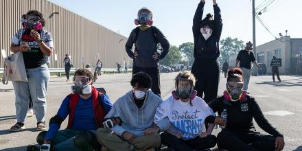 Protestors wearing gas masks sit on the ground with linked arms outside a big warehouse-facility. One person wears a t-shirt reading 'protect immigrants' and another, standing, holds up a US passport
