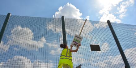 A woman in a high vis jacket is pictured from below standing next to a tall green fence. She holds a sign upside down that reads '#set her free'