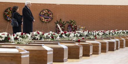 A man and a woman dressed in black stand solemnly by dozens of rows of wooden coffins laid out in a sports hall