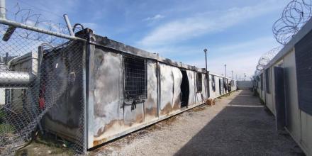 A burnt container is in the centre of the shot with a fence topped with barbed wire to the left. Containers without visible fire damage stand next to it further away, and a figure can be seen going into one