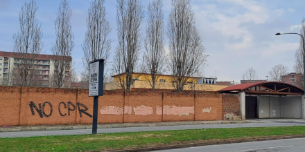 A brick wall with institutional-looking buildings behind it. On the wall is written 'No CPR' (Italian for 'no detention')