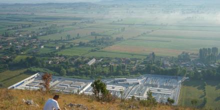 A person sits on a hill overlooking the detention centre in Gjader, Albania: a huge white sprawl of containers and buildings, contrasted against the green of the landscape around.