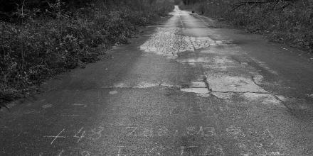 A black and white image of a road at the border of Croatia and Bosnia-Herzegovina. Two phone numbers are written on the road in white chalk – contact numbers for the Balkan route