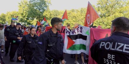 Protesters at Berlin's queer pride march walk along a street behind a big banner with the word 'Dyke' and a Palestine flag visible. In the foreground, a line of police officers walks alongside them 