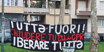 A banner strung between two trees, in front of a block of flats reads in Italian: "Tutta fuori! Chiudere Tutti i CPR. Iberare Tutta" / “Everyone out. Close all CPRs. Free them all”