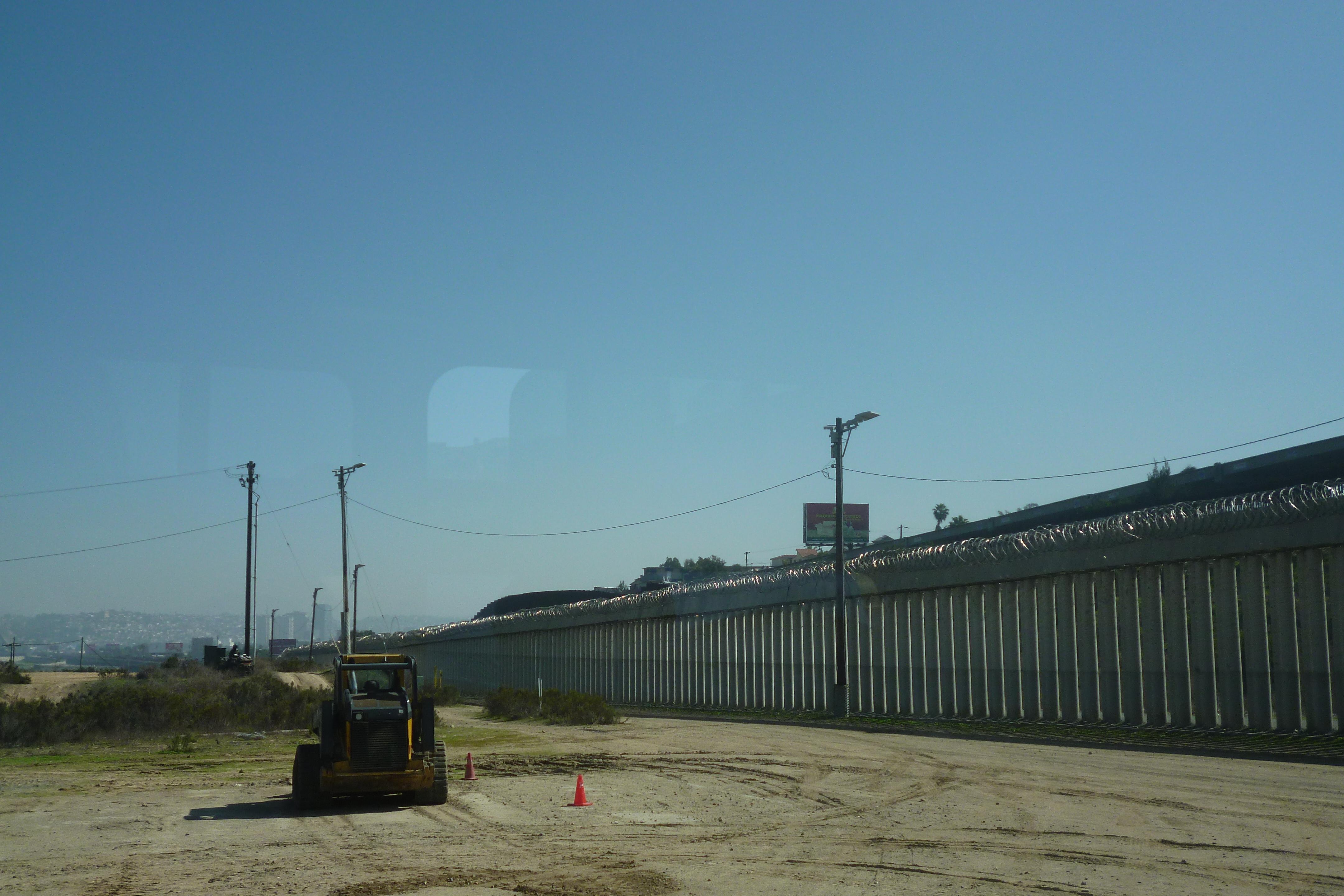 The picture shows a small tractor driving through a sandy field on a sunny day. On the right side of the picture you can see a long wall with barbed wire on top of it. In the background there are buildings of a city.