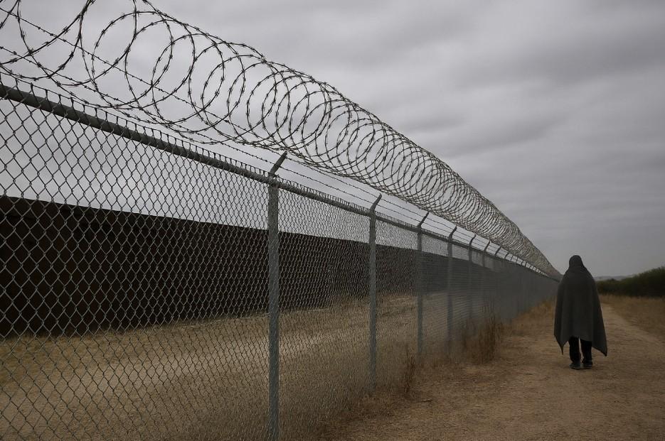 picture of a person seen from behind, walking next to a wired fence on a cloudy day