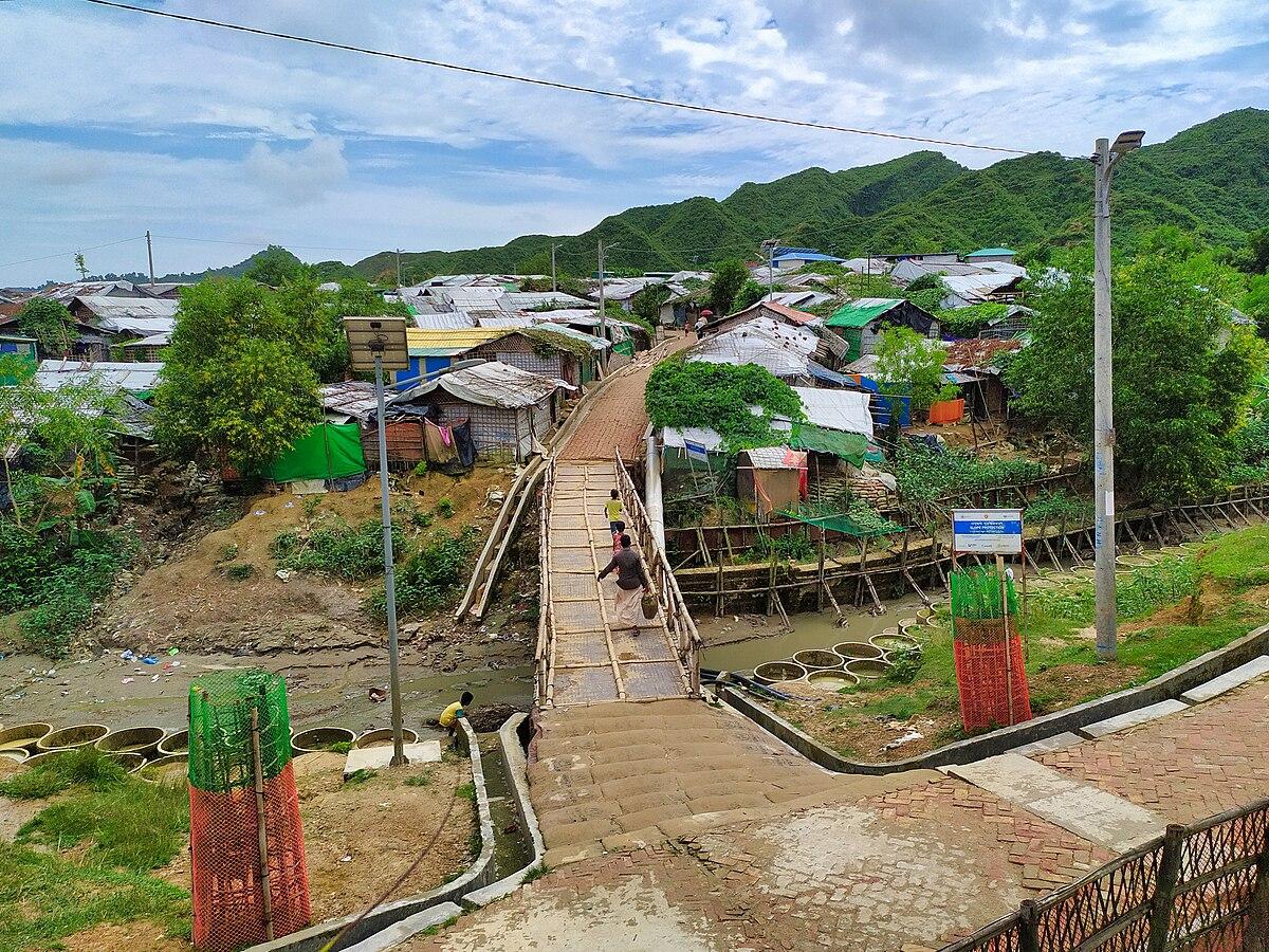 picture of a camp, with barracks in the back of the picture, and a wooden bridge in the middle, with people walking across it carrying bags of food