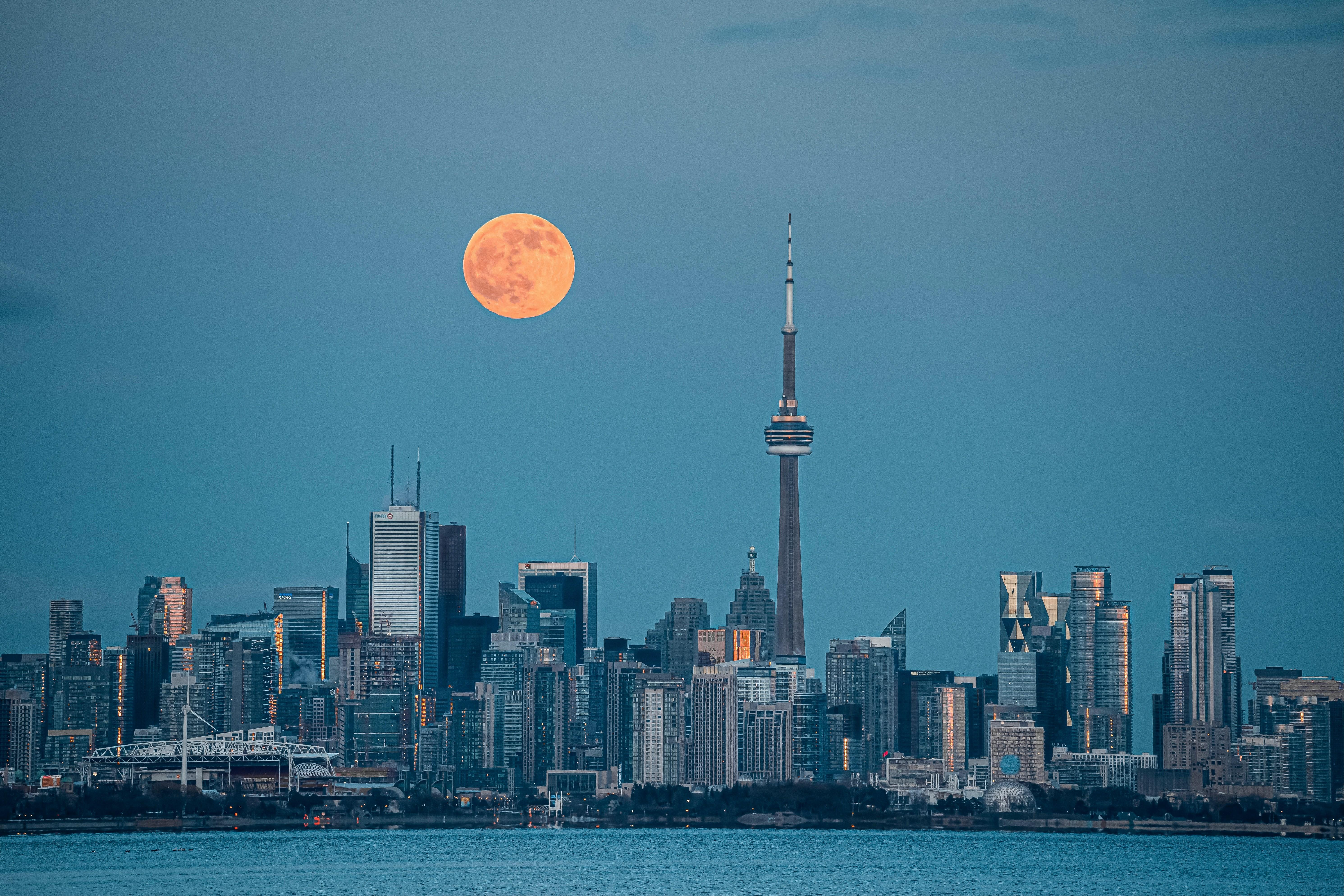 Toronto skyline as seen from the water. A full moon sits large and orange in the sky above the skyscrapers 
