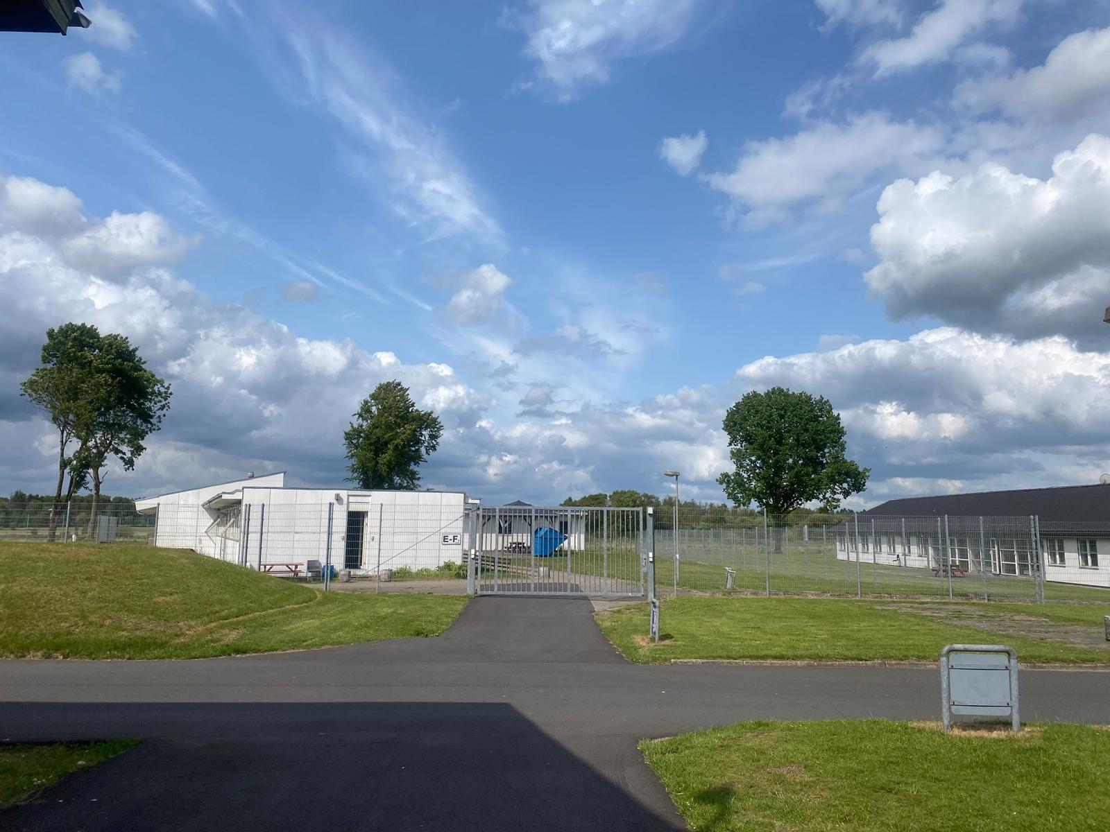 picture of a closed gate with a couple of white buildings behind it. In front of it, a small path crosses another with some green areas in between, and a blue sky.