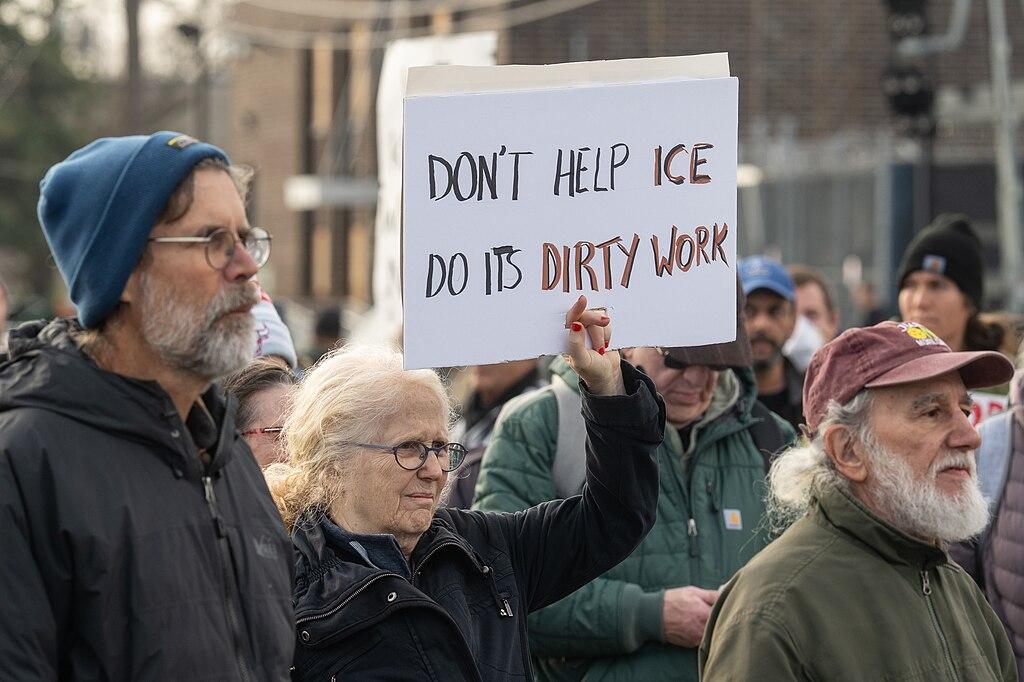 A woman holds up a sign reading 'Don't help ICE do its dirty work' standing beside other protesters 