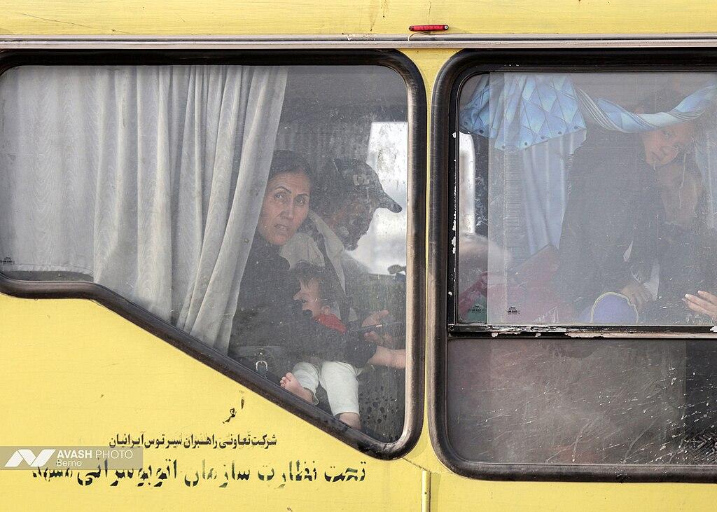 A woman sits next to a man –&nbsp;her and several children look out from the window of the yellow bus they are sat inside, with writing in Farsi on the outside of the bus